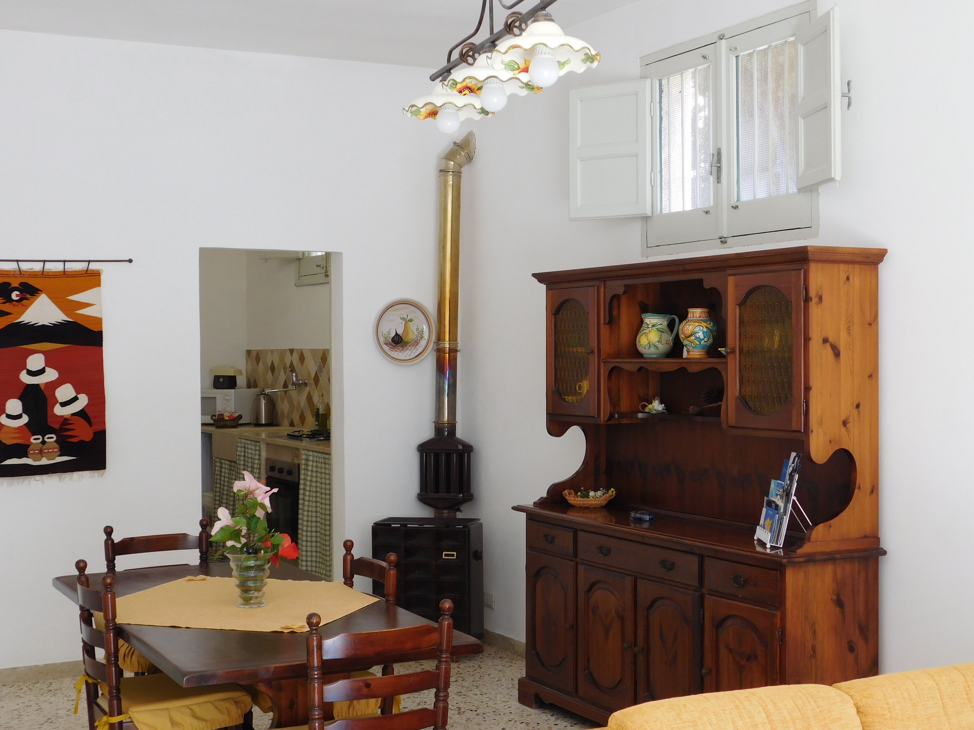A dining area with a wooden table, chairs, a vase with flowers, a decorative lamp, a stove, and a wooden cabinet with pottery.