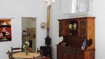 A dining area with a wooden table, chairs, a vase with flowers, a decorative lamp, a stove, and a wooden cabinet with pottery.