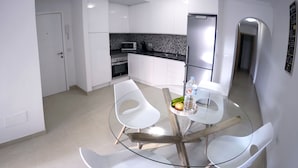 Une cuisine moderne avec des meubles blancs, un four à micro-ondes, un réfrigérateur et un coin repas comprenant une table en verre et des chaises blanches.