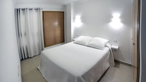 Un lit bien fait dans une pièce où se trouvent une armoire en bois, un rideau blanc et deux lampes fixées au mur.