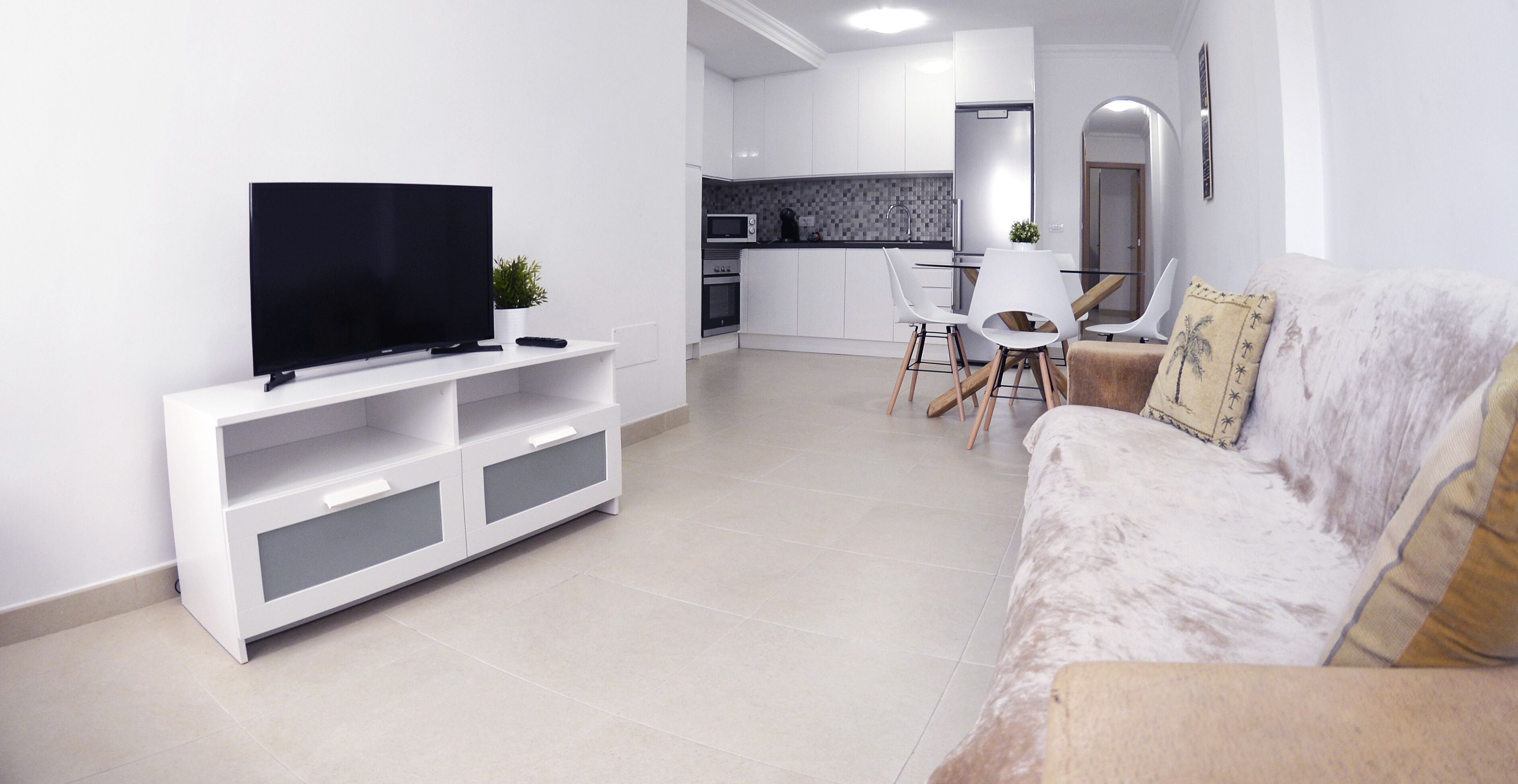Un salon moderne avec un téléviseur à écran plat posé sur un meuble de rangement blanc, un canapé beige agrémenté de coussins décoratifs et un coin repas avec des chaises blanches et une table en verre.