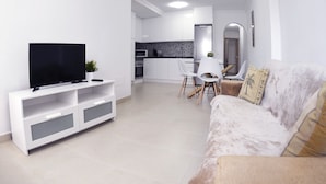 Un salon moderne avec un téléviseur à écran plat posé sur un meuble de rangement blanc, un canapé beige agrémenté de coussins décoratifs et un coin repas avec des chaises blanches et une table en verre.