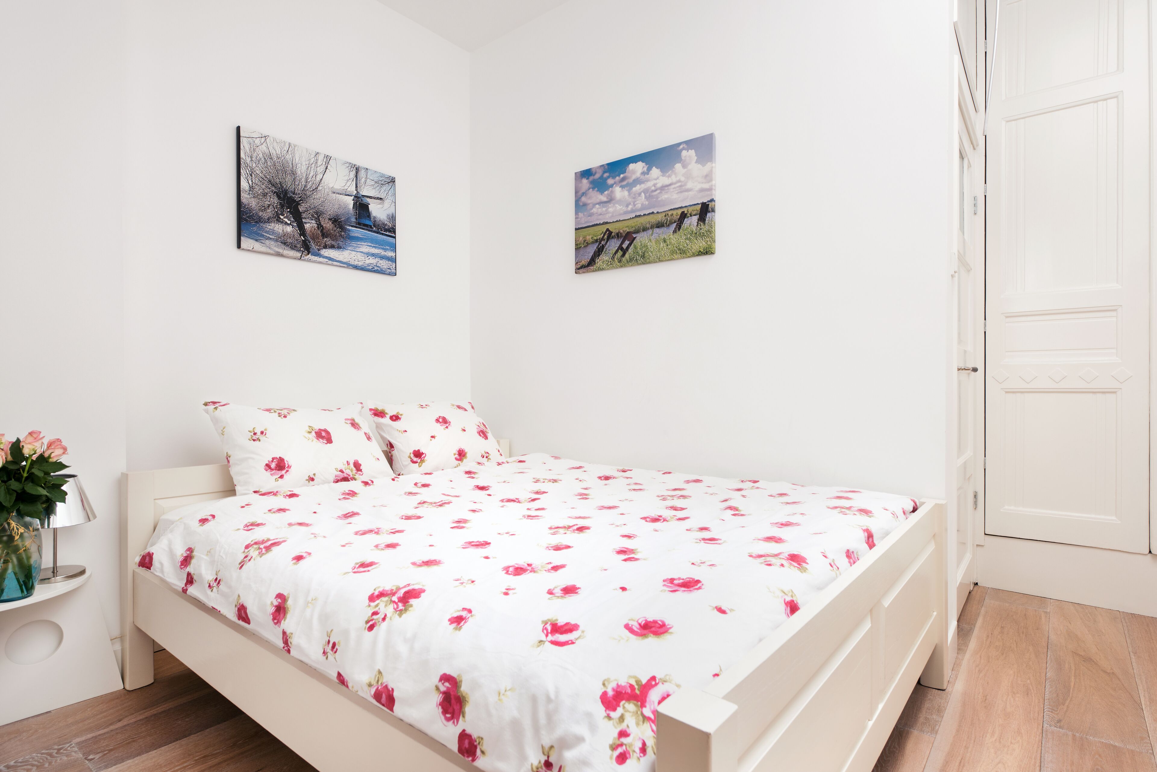 Ein Schlafzimmer mit einem Bett, einem Nachttisch, einer Blumenvase und zwei gerahmten Bildern an der Wand.