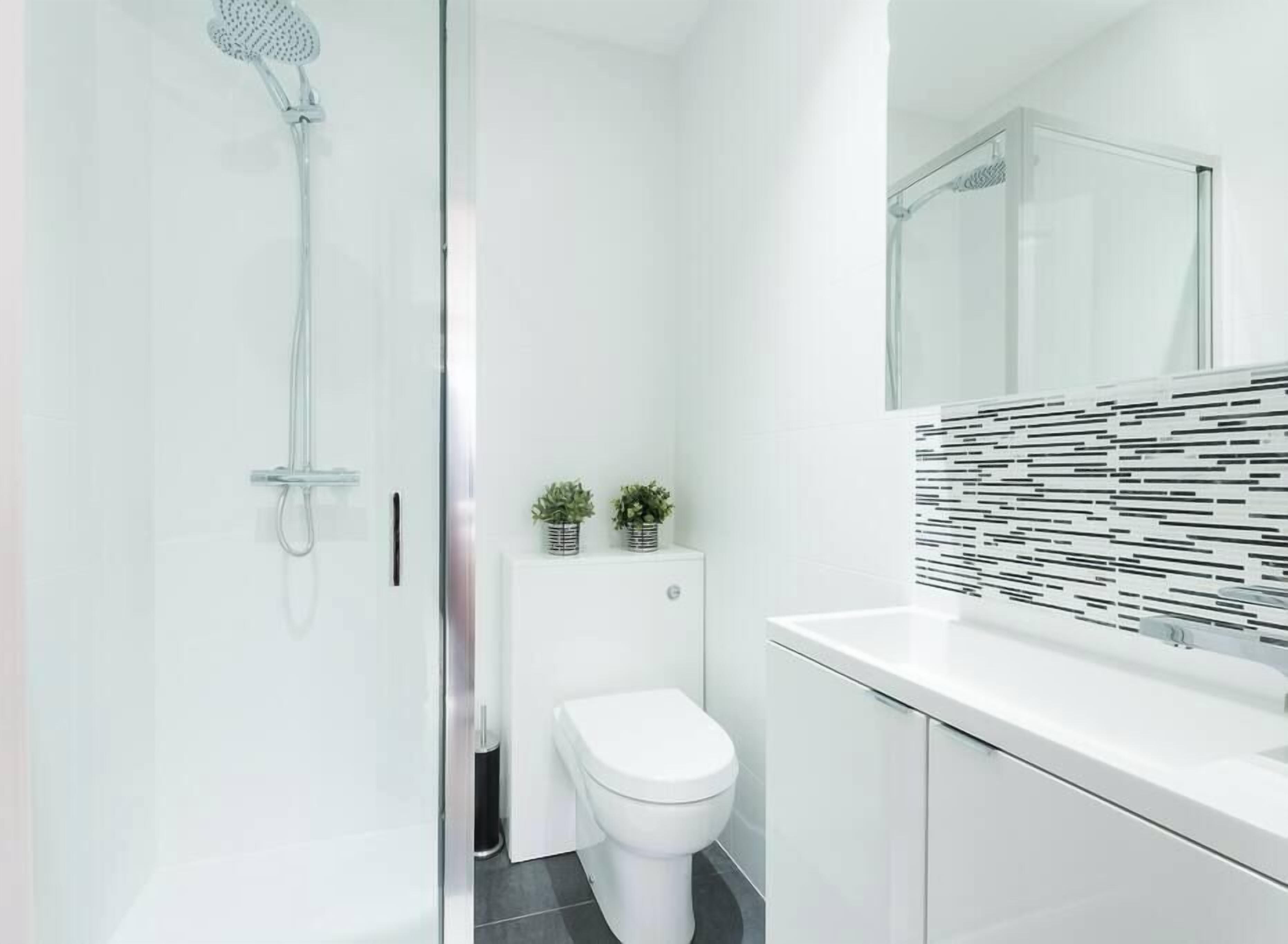 Une salle de bain moderne avec une douche à l’italienne en verre, des toilettes blanches et un lavabo blanc avec un pare-éclaboussures carrelé en noir et blanc.