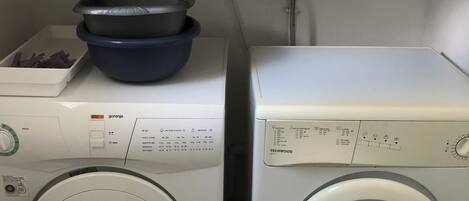 A laundry area with two washing machines, a shelf with folded towels, and a container with cleaning supplies.