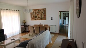 A living room with a sofa, a coffee table, and a dining area with a table and chairs. There is a painting on the wall and a kitchen visible in the background. - Farmhouse with large outdoor garden, private, near the beach, very green and quiet (Alfeizerão - São MArtinho do porto)
