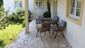 A covered patio with stone flooring, wooden chairs with patterned cushions, and a view of a house with a tiled roof. - Farmhouse with large outdoor garden, private, near the beach, very green and quiet (Alfeizerão - São MArtinho do porto)