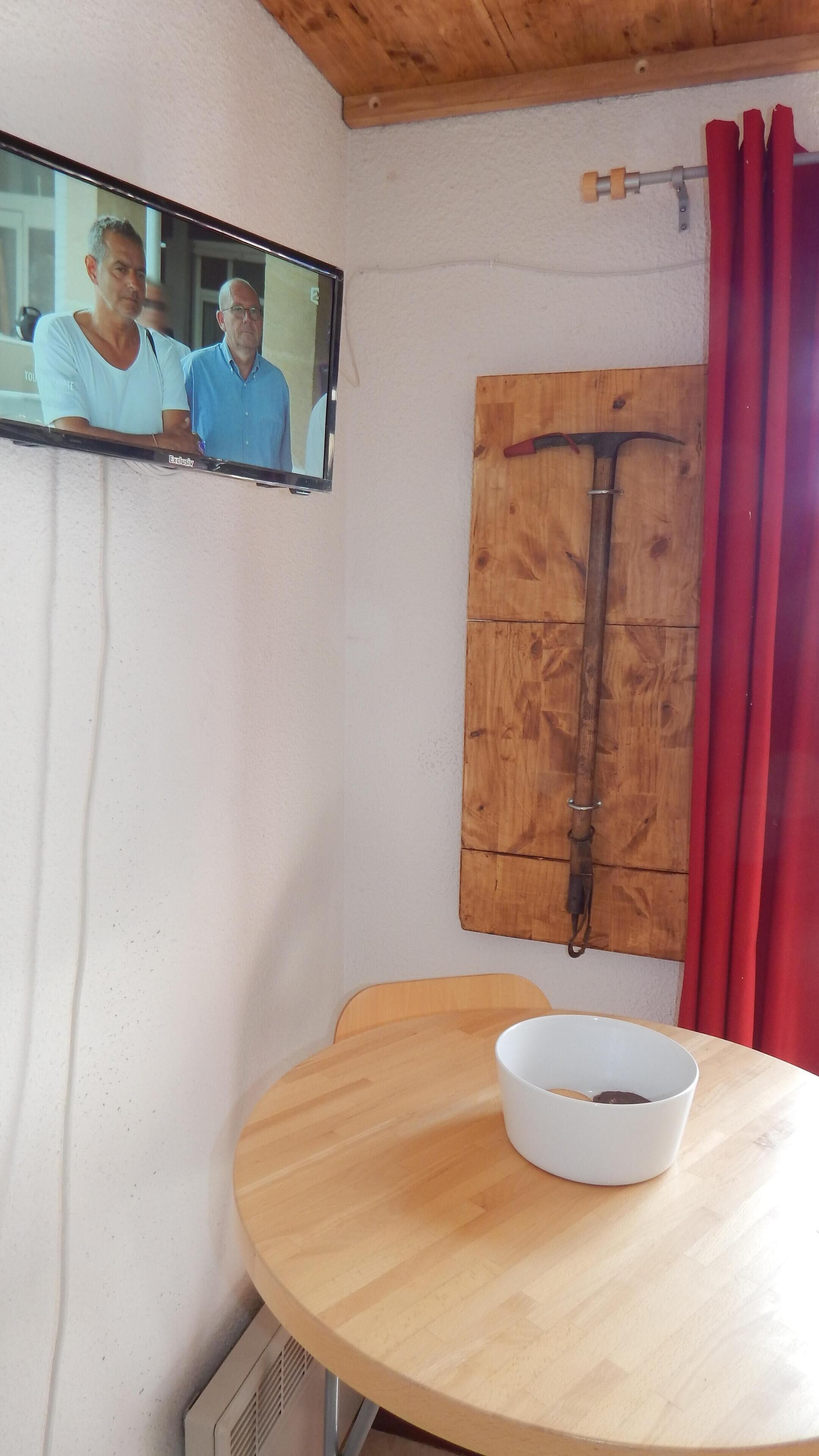 Une pièce avec une table en bois, un bol blanc, un téléviseur fixé au mur et un volet en bois.