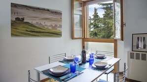 Un coin repas avec une table dressée pour quatre personnes, une grande fenêtre donnant sur un paysage arboré et un tableau représentant un paysage accroché au mur.