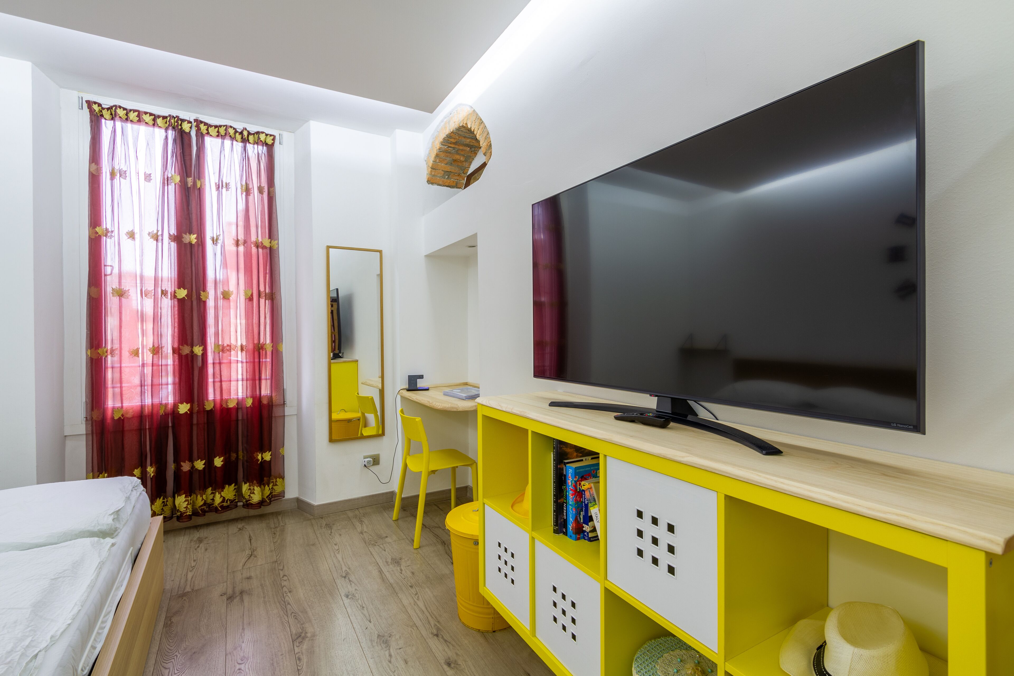 Een slaapkamer met een bed, een bureau, een televisie en een geel kastje.
