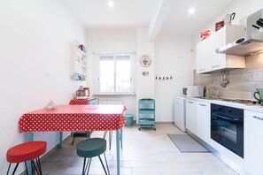 A compact kitchen with white cabinetry, a red and white polka dot table, teal stools, and a window with blinds.