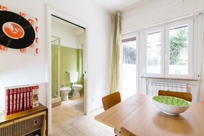 A room with a wooden table, a bowl, a radiator, a window with a view, and a door leading to a bathroom.