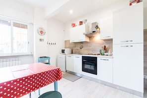 Een moderne keuken met witte kasten, een magnetron, een vaatwasser en een fornuis. Er is een raam met zonwering, een tafel met een tafelkleed met stippen, en een klein keukeneiland.