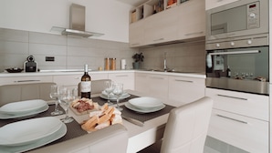 A modern kitchen with white cabinetry, a built-in oven, and a dining area set for four with plates, glasses, and a bottle of wine.