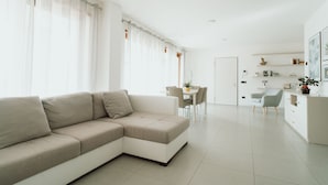 A modern living room with a beige sectional sofa, a dining area with a table and chairs, and a kitchen area with white cabinets and a countertop.