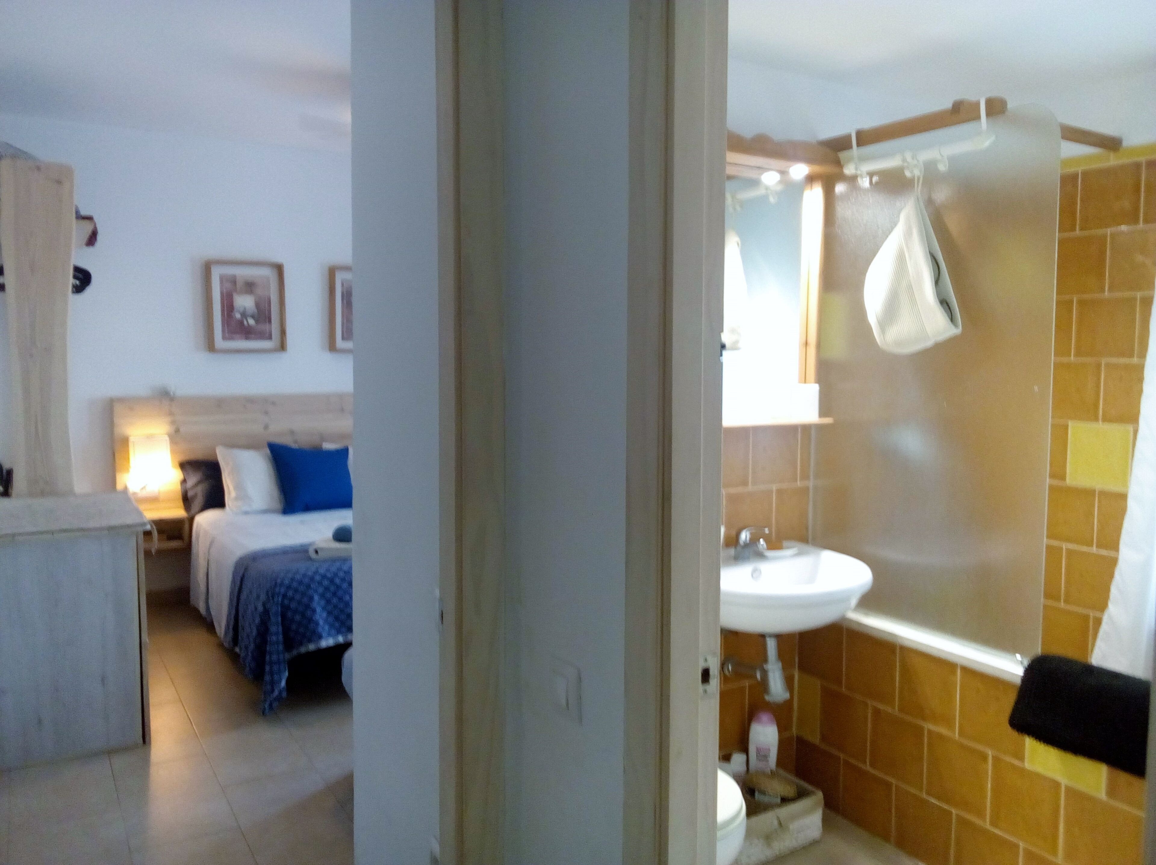 A bedroom with a bed, bedside table, and a nightstand. There is a bathroom with a sink, mirror, and towel rack.