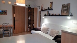 Um quarto aconchegante com uma cama, mesa de cabeceira e escrivaninha com cadeira. Há uma estante e uma planta pendente.
