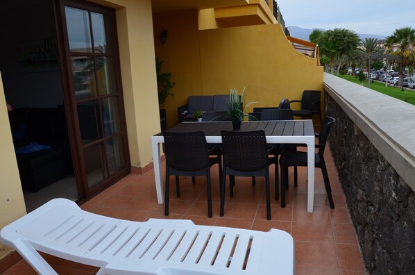 An outdoor patio with a table and chairs, a white lounge chair, and a yellow wall.