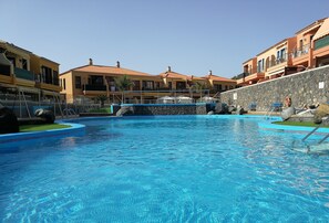 A swimming pool with clear blue water, surrounded by buildings with orange and yellow facades, and a stone wall.