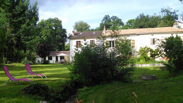 A white building with a tiled roof, surrounded by lush greenery and a small stream. There are two purple lounge chairs in the foreground. - At the water wire Perigord (Lisle)