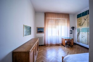 A bedroom with a wooden floor, a bed, a desk with a chair, a radiator, and a window with sheer curtains.