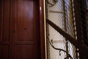 Una puerta de madera con manija y placa de latón, que da paso a una habitación con pared estampada y baranda metálica.