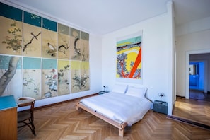 A bedroom with a wooden floor, a bed, a desk, and a chair. There are multiple wall panels with artwork.