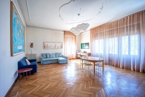 A spacious living room with a herringbone wooden floor, a blue sofa, a dining table with chairs, and large windows with sheer curtains.