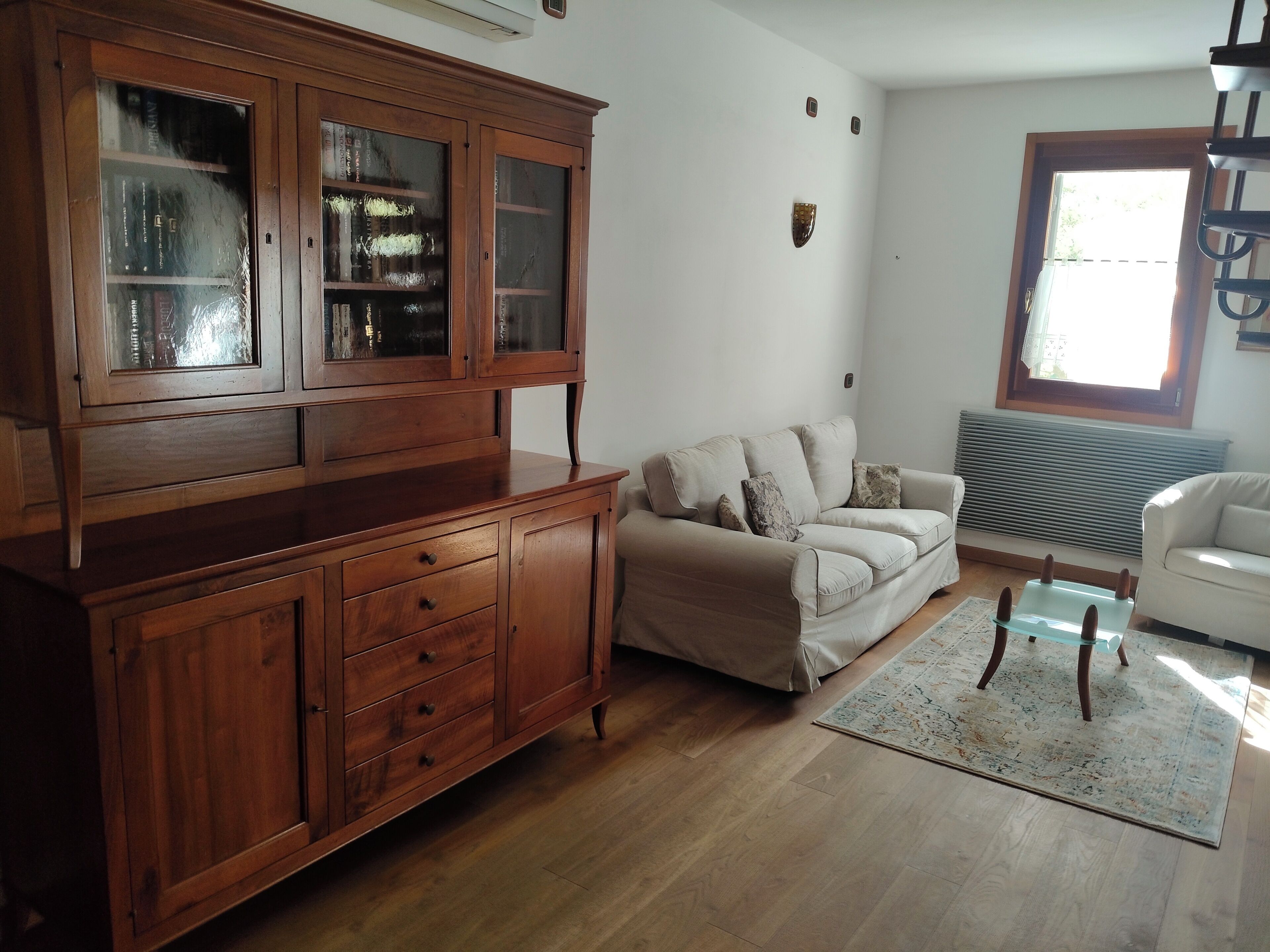 A living room with a wooden cabinet, a beige sofa, a glass coffee table, and a white armchair.