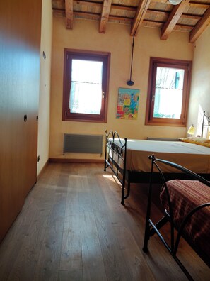 Ein Zimmer mit Holzböden, einem Bett mit Metallgestell, zwei Fenstern mit Holzrahmen, einem Heizkörper und einer Wandleuchte.