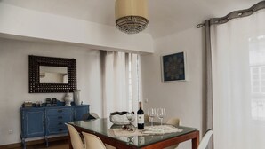 A dining area with a glass table, two chairs, a blue cabinet, a chandelier, and a window with curtains. - Casa Resende - Resende House (Évora)