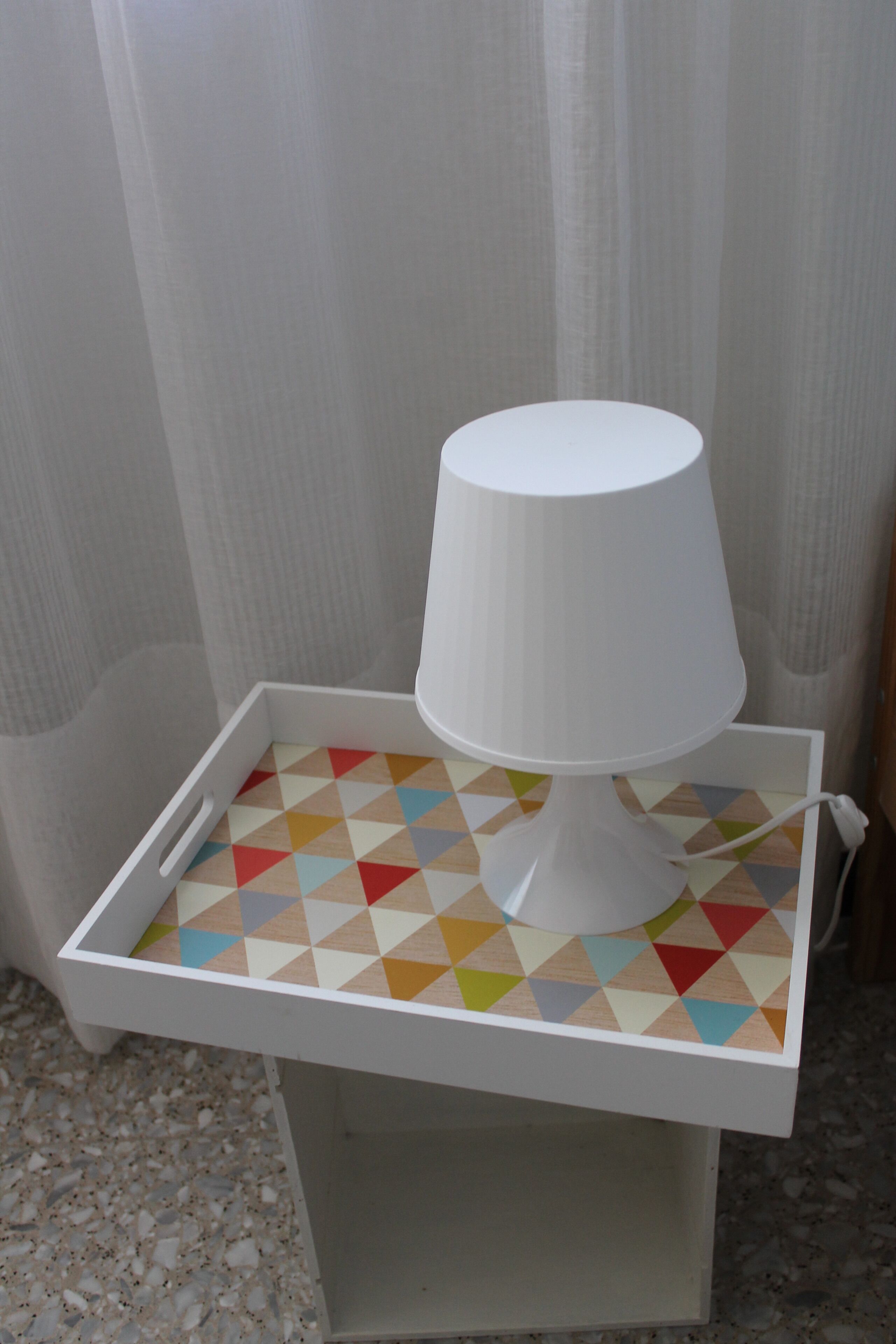 A white bedside table with a lamp, a geometric-patterned tray, and a small drawer.
