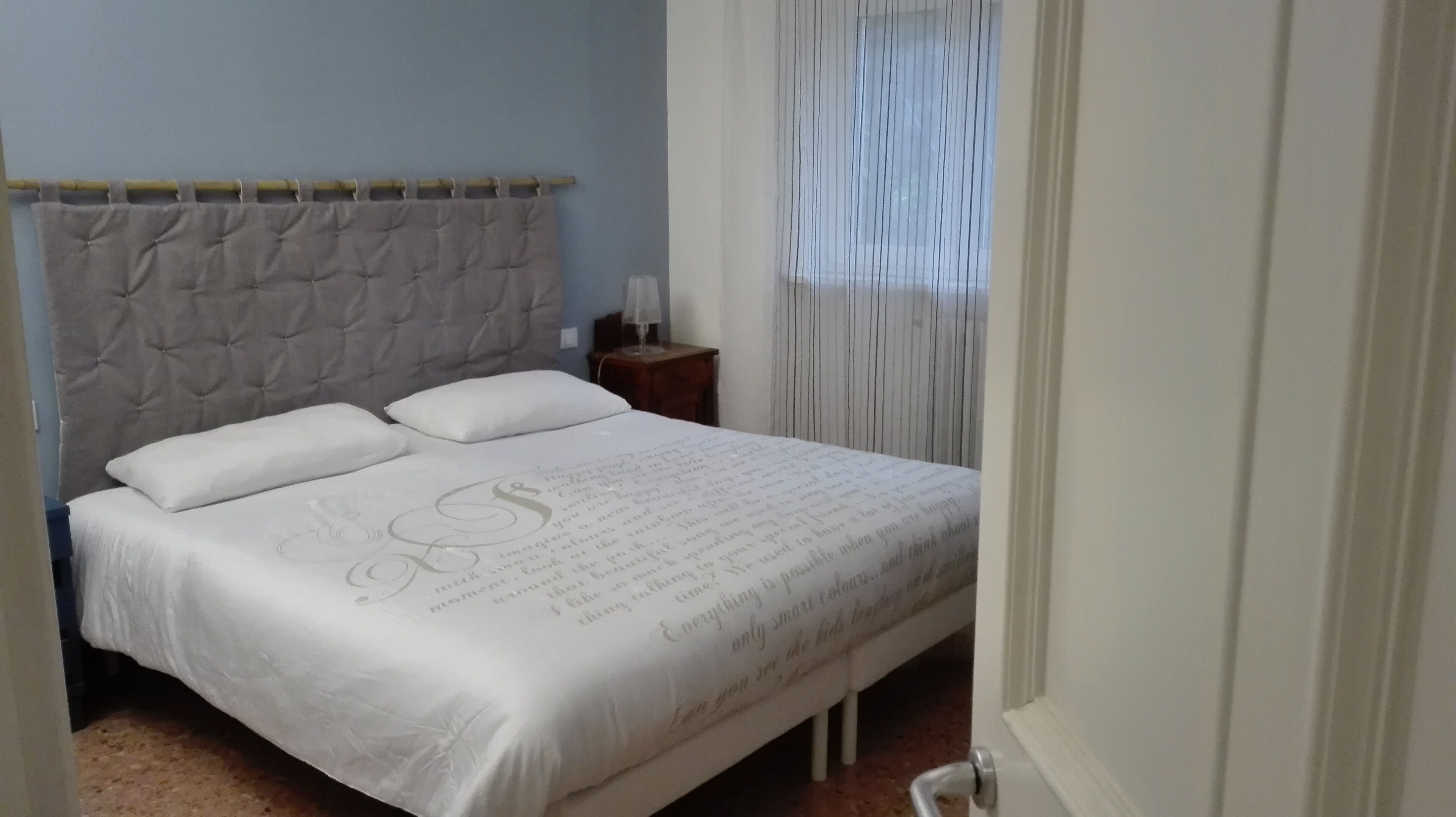 A neatly made bed with a tufted headboard, a bedside table with a lamp, and a window with vertical blinds.