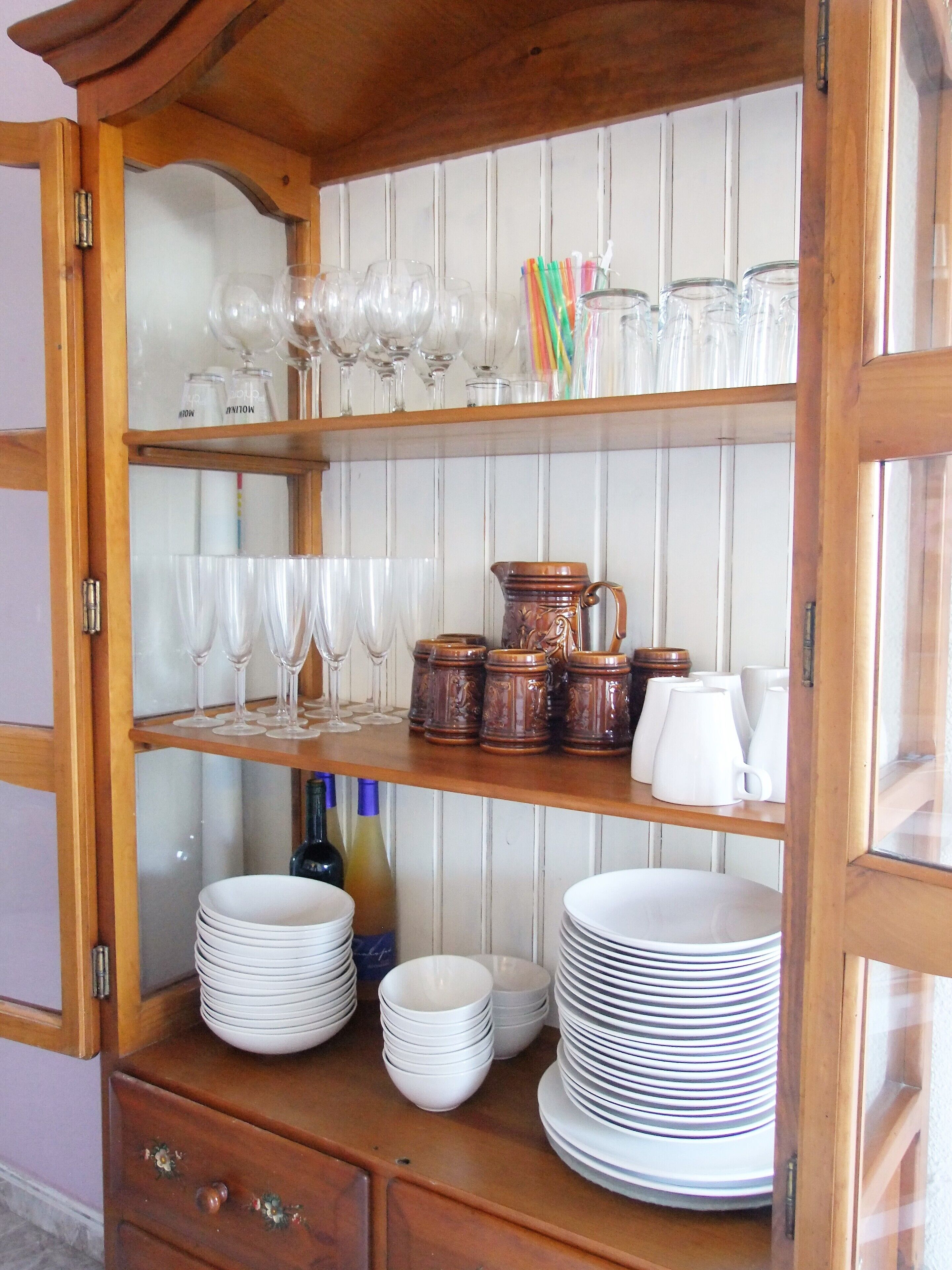 A wooden cabinet with multiple shelves containing glassware, ceramic mugs, and stacked plates.