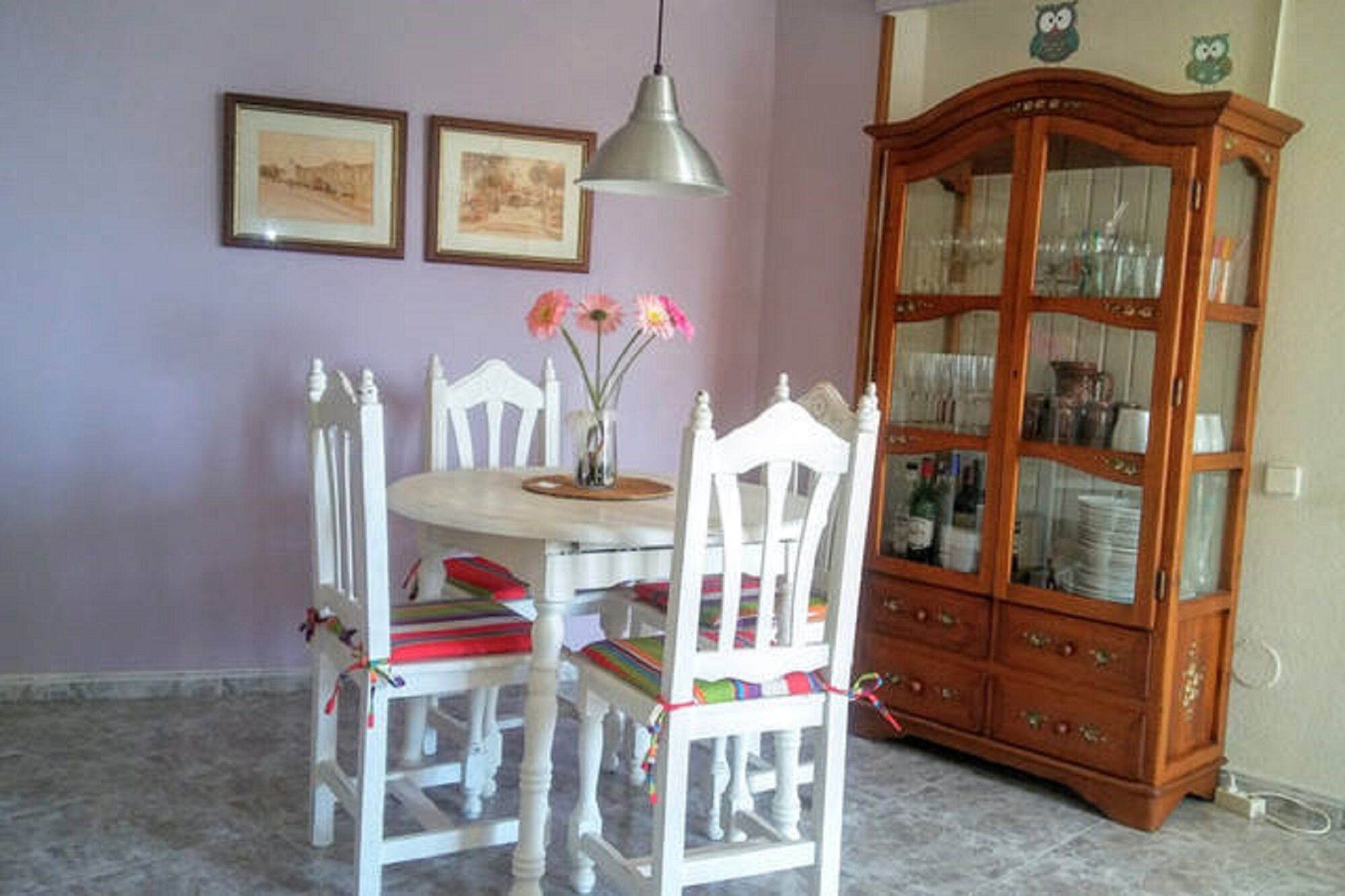 A dining area with a round table, white chairs, a vase with flowers, and a wooden cabinet with glass doors.