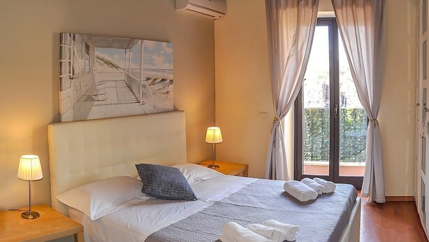 A hotel room with a bed, two bedside lamps, a painting, an air conditioning unit, and a balcony with curtains. - Bellini Apartment with Balcony   (Catania)