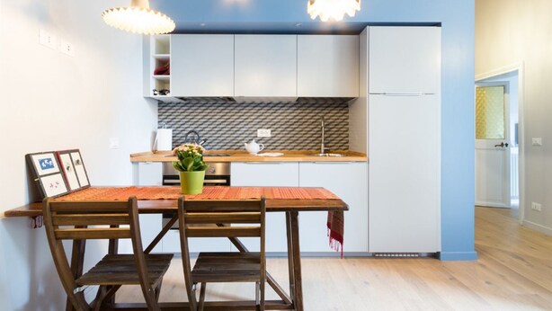 Een kleine keuken met witte kastjes, een houten aanrechtblad en een eethoek met een houten tafel en stoelen.