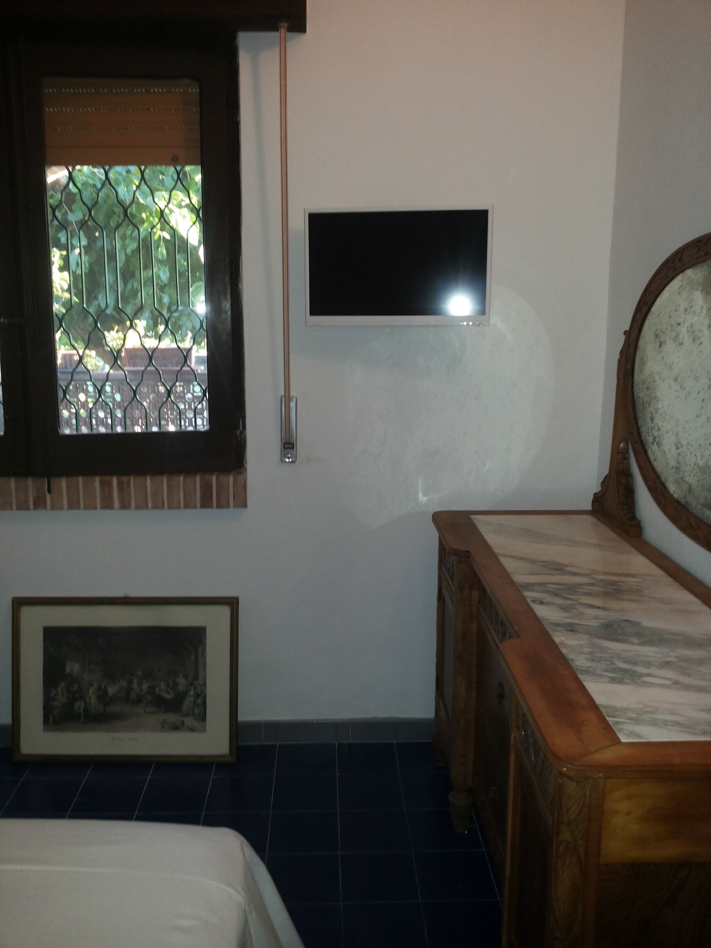 Une chambre à coucher avec un lit, un téléviseur fixé au mur, une fenêtre avec une grille en métal et un tableau encadré accroché au mur.