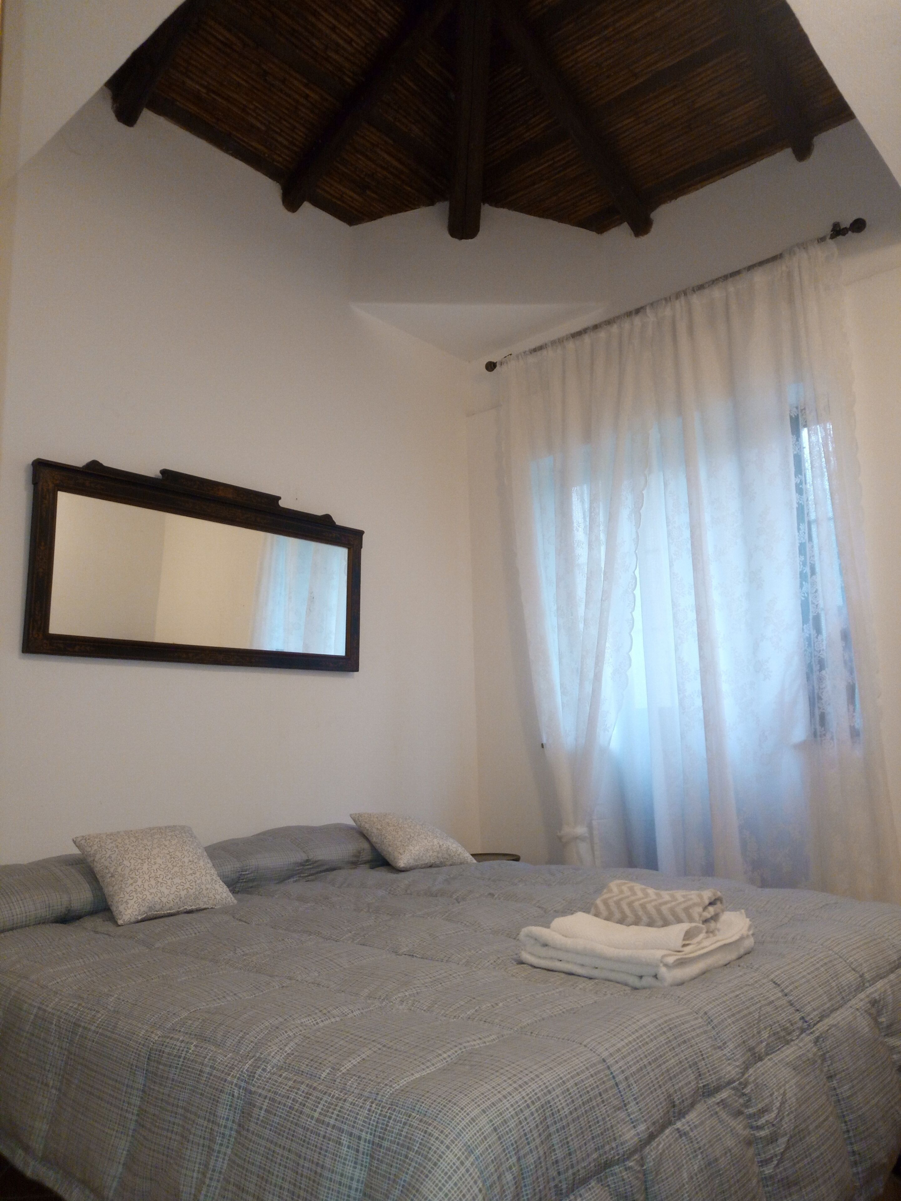 A bedroom with a bed, a mirror, and a window with sheer curtains.