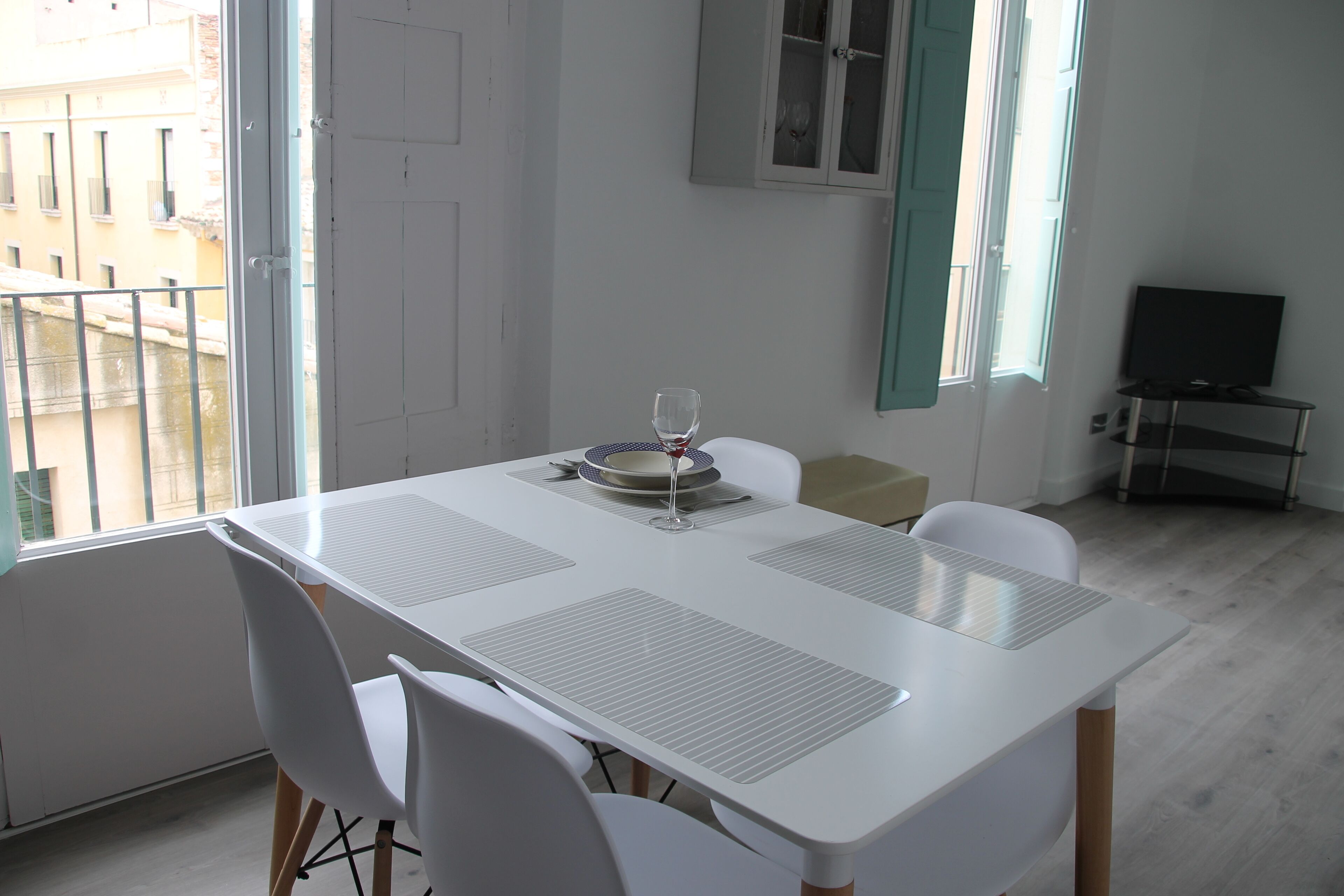 Zona pranzo con tavolo e sedie bianche, un bicchiere di vino e vista sugli edifici esterni.