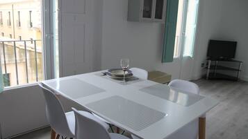 Zona pranzo con tavolo e sedie bianche, un bicchiere di vino e vista sugli edifici esterni.