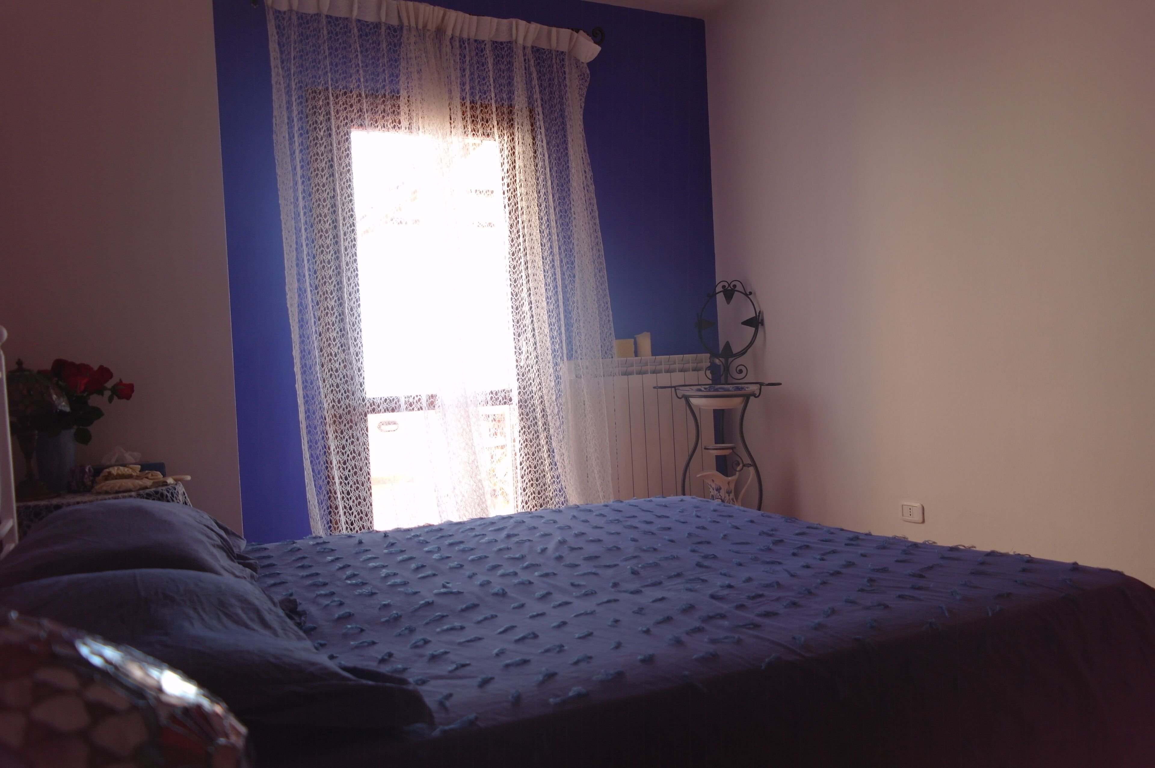 A bedroom with a bed, a window with lace curtains, a bedside table with a mirror, and a wall clock.
