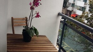 Un balcón con una mesa y sillas de madera, una planta en maceta con flores rosadas, y vista a un edificio moderno al otro lado de la calle.