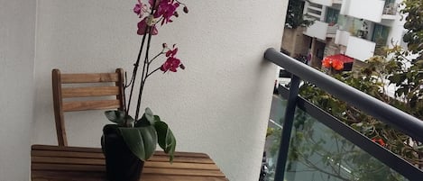Un balcón con una mesa y sillas de madera, una planta en maceta con flores rosadas, y vista a un edificio moderno al otro lado de la calle.