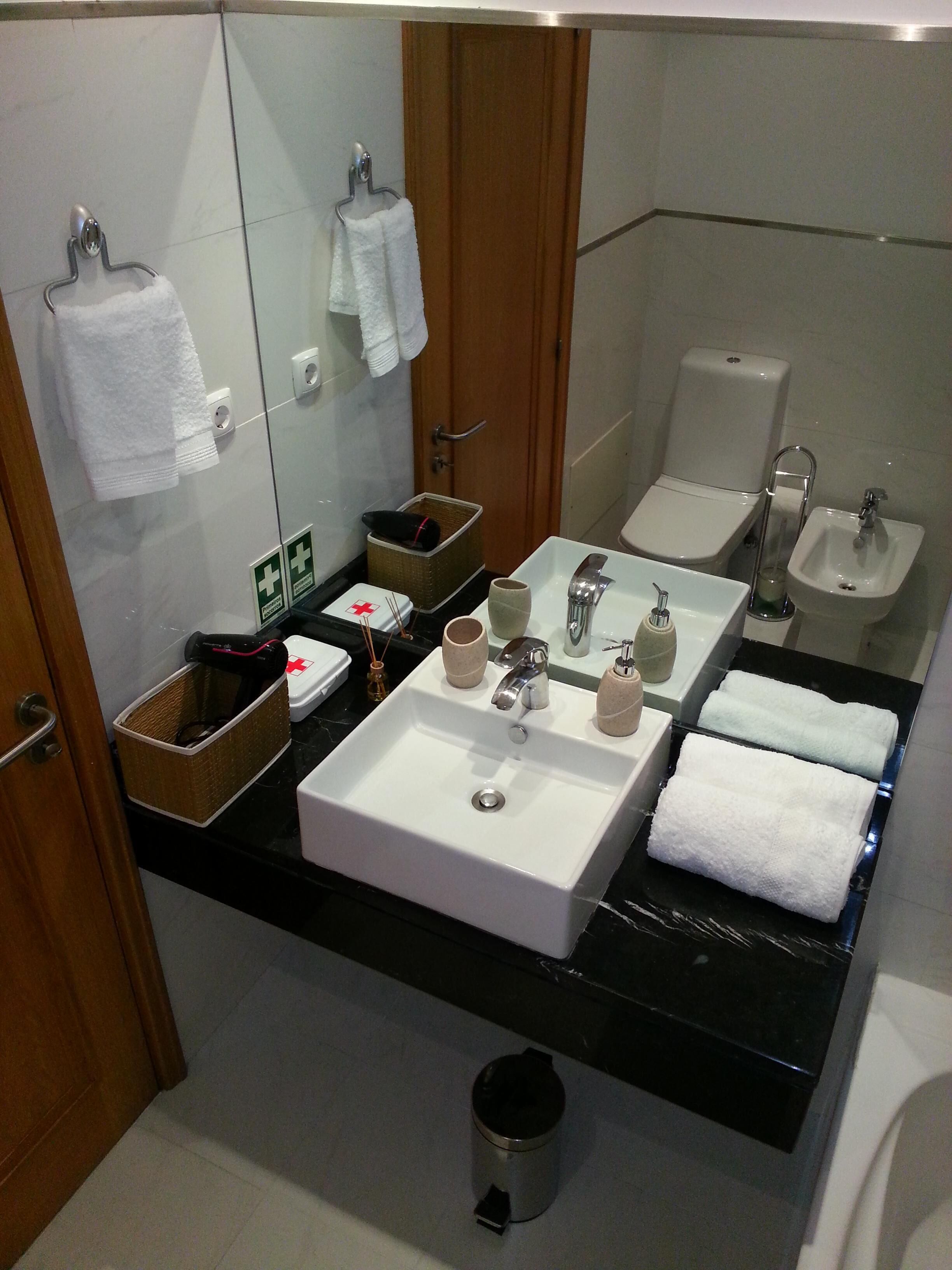 A modern bathroom with a large sink, a bidet, and a mirror. There are towels hanging on the wall and a trash can on the floor.