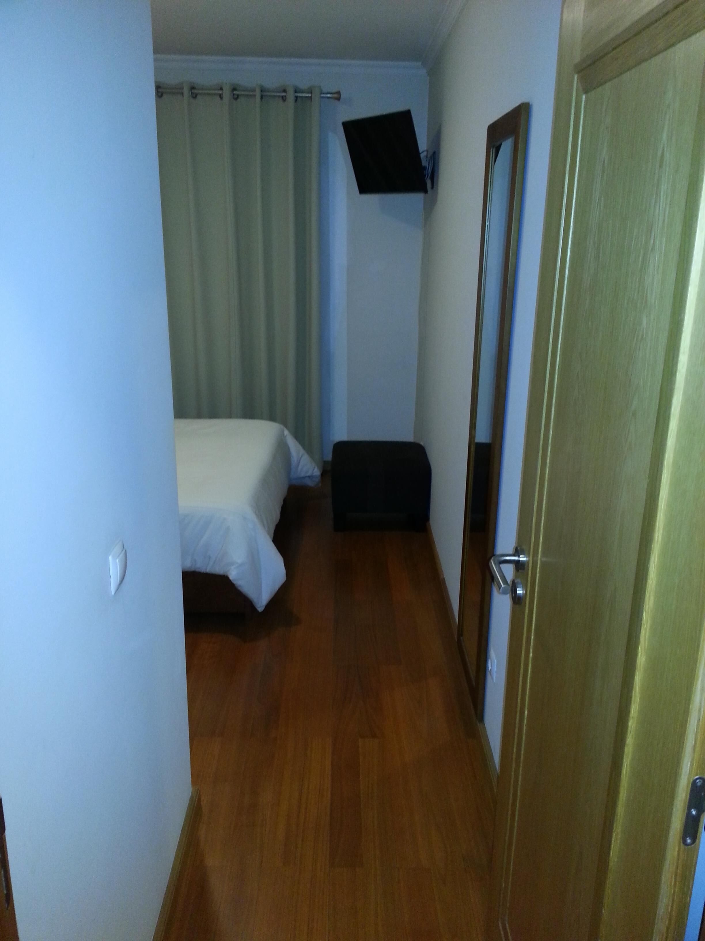 A small, narrow room with a bed, a wall-mounted TV, a door, and a mirror.