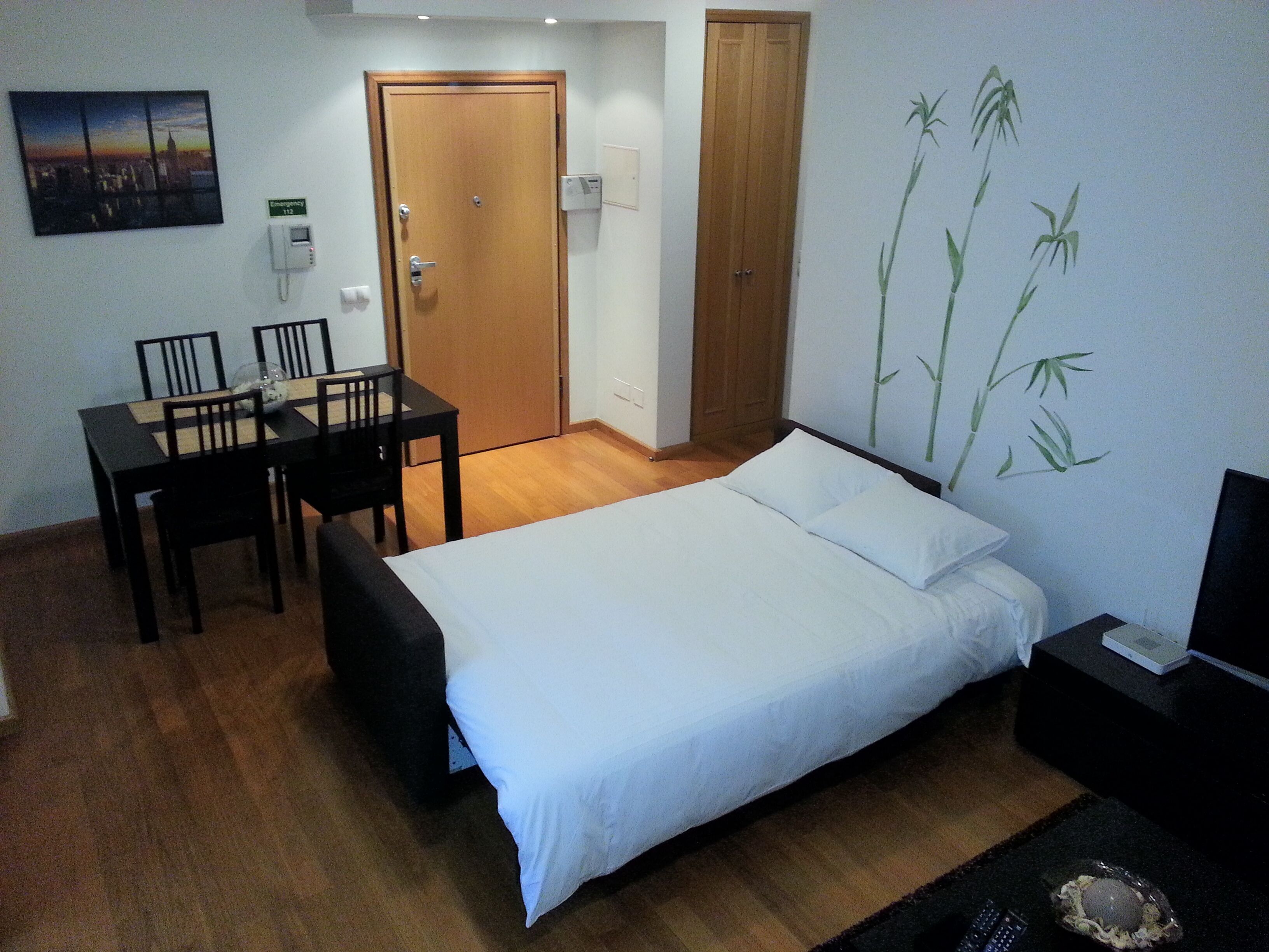 A bedroom with a bed, a desk with chairs, a wooden wardrobe, and a wall-mounted emergency phone.