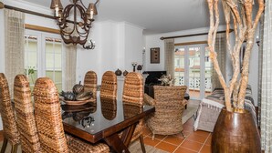 A dining area with a glass table surrounded by wicker chairs, a chandelier, and a view of a balcony with a railing. - Apartment in Praia D'El Rey Golf & Beach Resort (Amoreira)
