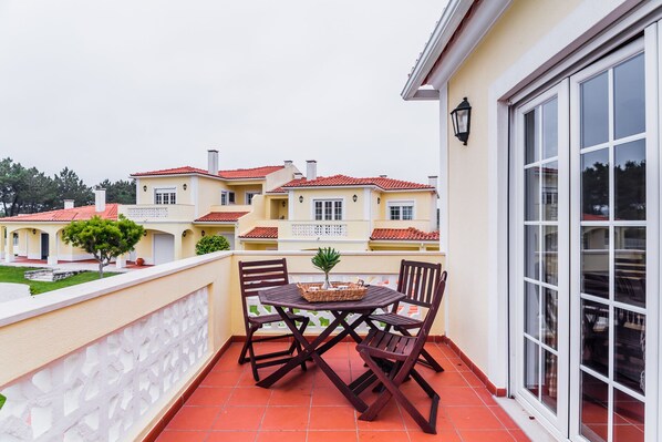 Un balcon avec une table et des chaises, un pot de fleurs et une vue sur un immeuble jaune aux toits rouges.