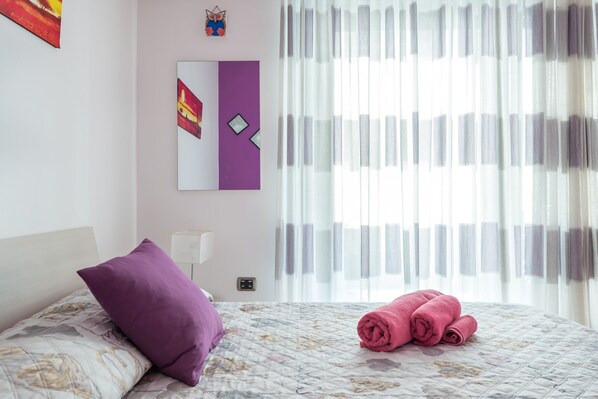 A hotel room with a bed, a purple pillow, rolled towels, a wall-mounted mirror, and a window with sheer curtains. - The Myth. Central in Ortigia. Kid-friendly (Siracusa)